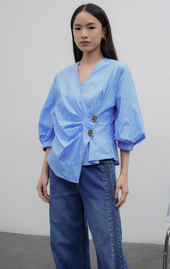 Blus wanita motif garis warna biru (blue) Castell Top berbahan cotton-polyester blend dengan kerah V, aksen kerutan di pinggang samping (side-waist ruching), kancing dekoratif, dan lengan 3/4 bermodel balon.