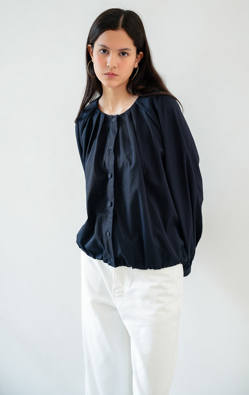 Atasan wanita lengan panjang warna biru navy (navy blue) Catharine Top berbahan katun twill (cotton twill) dengan kerah bulat, bukaan kancing depan, dan detail tali serut di bagian bawah (drawstring hem). 