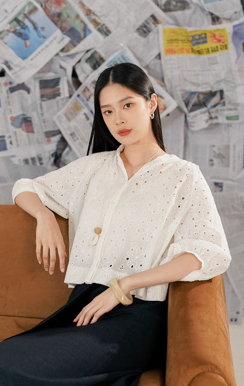 Model wanita mengenakan Ceara Top White, sebuah atasan blouse putih bahan crochet/eyelet dengan motif bunga berlubang. Baju memiliki kancing depan, kerah V rendah tanpa lapel, dan lengan 3/4 model balon (puff sleeves) untuk gaya vintage modern.