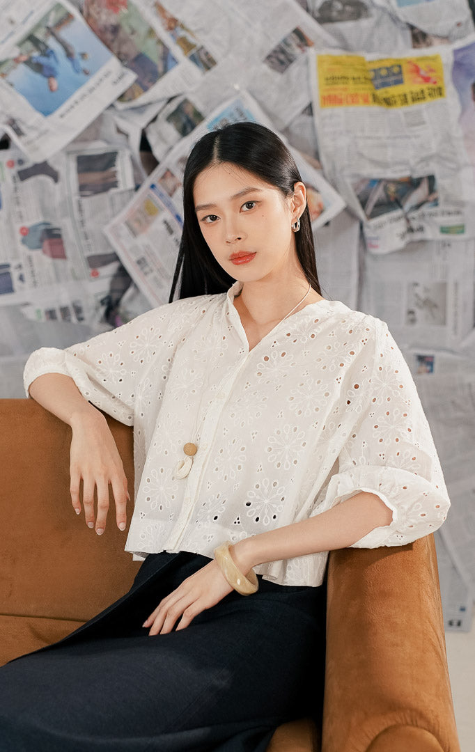 Model wanita mengenakan Ceara Top White, sebuah atasan blouse putih bahan crochet/eyelet dengan motif bunga berlubang. Baju memiliki kancing depan, kerah V rendah tanpa lapel, dan lengan 3/4 model balon (puff sleeves) untuk gaya vintage modern.