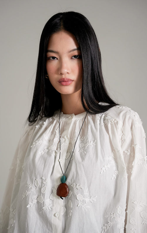 Colette Top warna putih berbahan katun dengan detail tekstur bordir bunga, dipadukan dengan celana jeans cutbray biru, kalung panjang, dan sepatu heels putih.