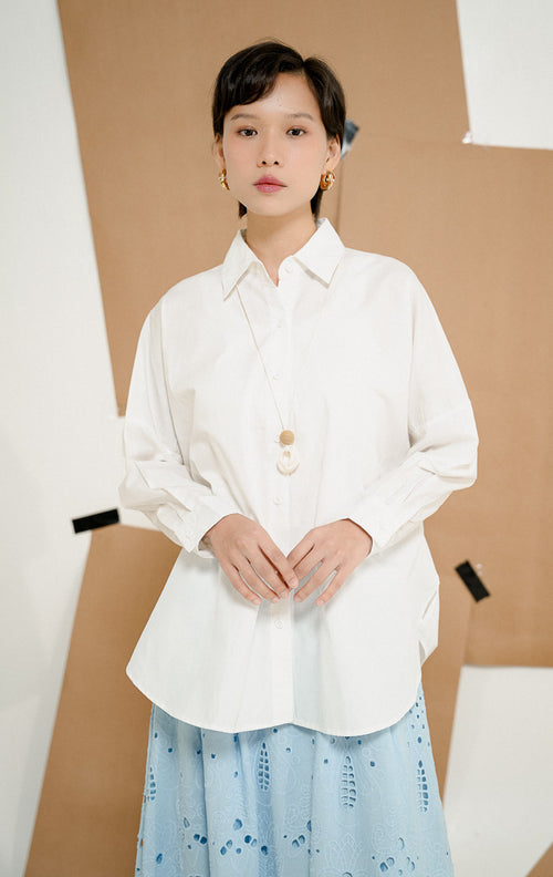 Model tampak samping mengenakan Dayle Top White, produk kemeja kerja kantor wanita lengan panjang warna putih dengan detail ruffle di punggung dipadukan rok panjang biru. 