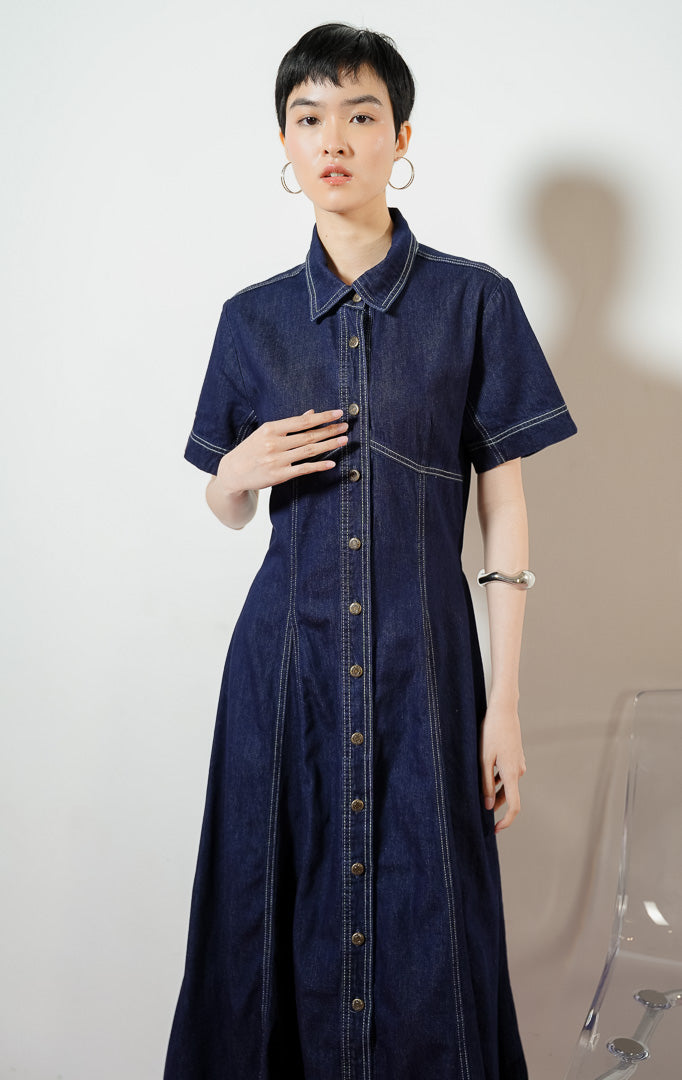 Model wanita mengenakan Lumera Classic Flared Maxi Dress warna dark denim, gaun dress panjang bahan jeans wanita lengan pendek berwarna biru dongker dengan detail jahitan kontras putih. 