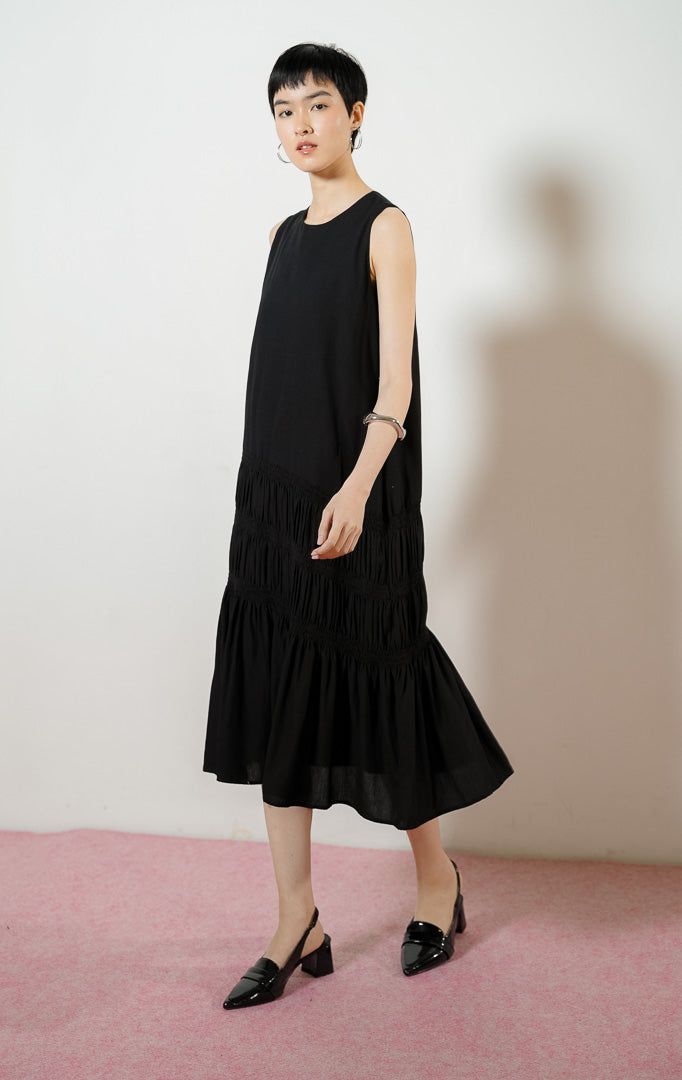 Model wanita mengenakan Ravena Shirred Bodice Drop-Tier Maxi Dress warna black, gaun panjang dress tanpa lengan wanita warna hitam dengan detail aksen serut karet smok bertingkat di rok. 