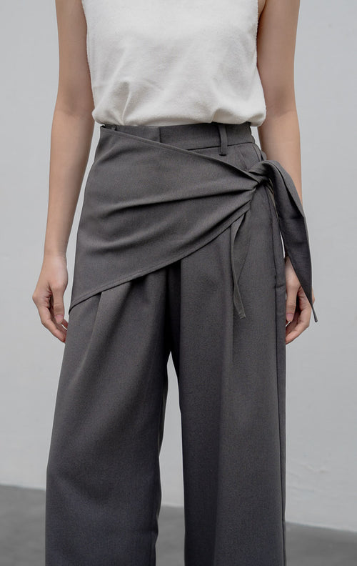 Celana kulot wanita model longgar (wide-leg pants) warna abu-abu (grey) Edliana Pants berbahan cotton-polyester dengan detail lapisan silang (overlap wrap), sabuk tali asimetris (asymmetrical belt), dan karet pinggang belakang.