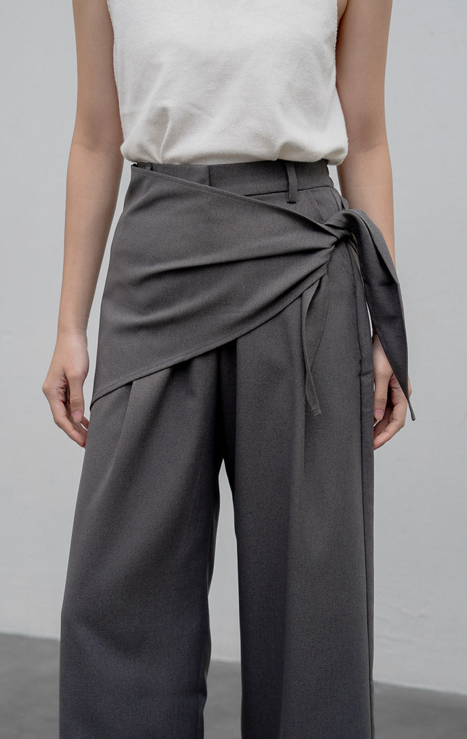 Celana kulot wanita model longgar (wide-leg pants) warna abu-abu (grey) Edliana Pants berbahan cotton-polyester dengan detail lapisan silang (overlap wrap), sabuk tali asimetris (asymmetrical belt), dan karet pinggang belakang.