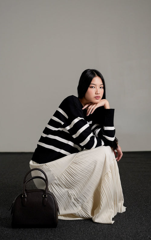 Elbie Striped Knit Sweater lengan panjang warna hitam dengan motif garis horizontal putih, dipadukan dengan rok panjang lipit warna krem dan sepatu mules hitam.