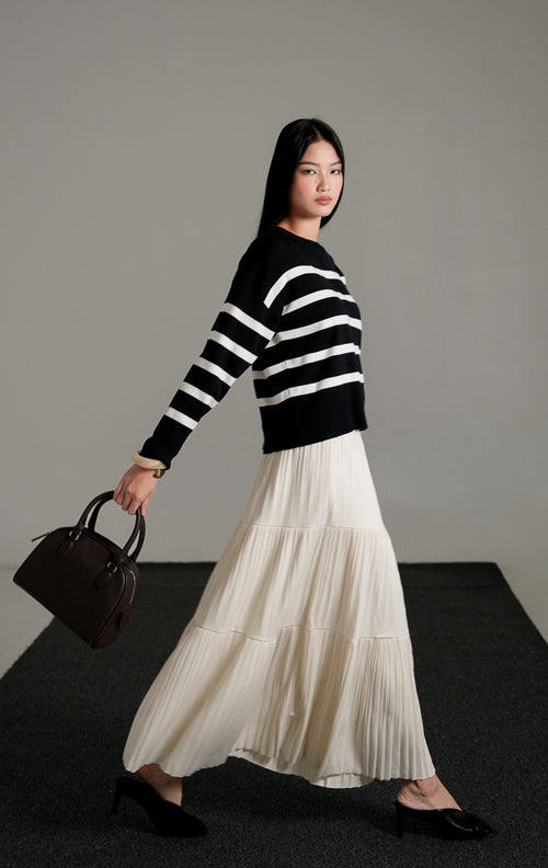 Elbie Striped Knit Sweater lengan panjang warna hitam dengan motif garis horizontal putih, dipadukan dengan rok panjang lipit warna krem dan sepatu mules hitam.