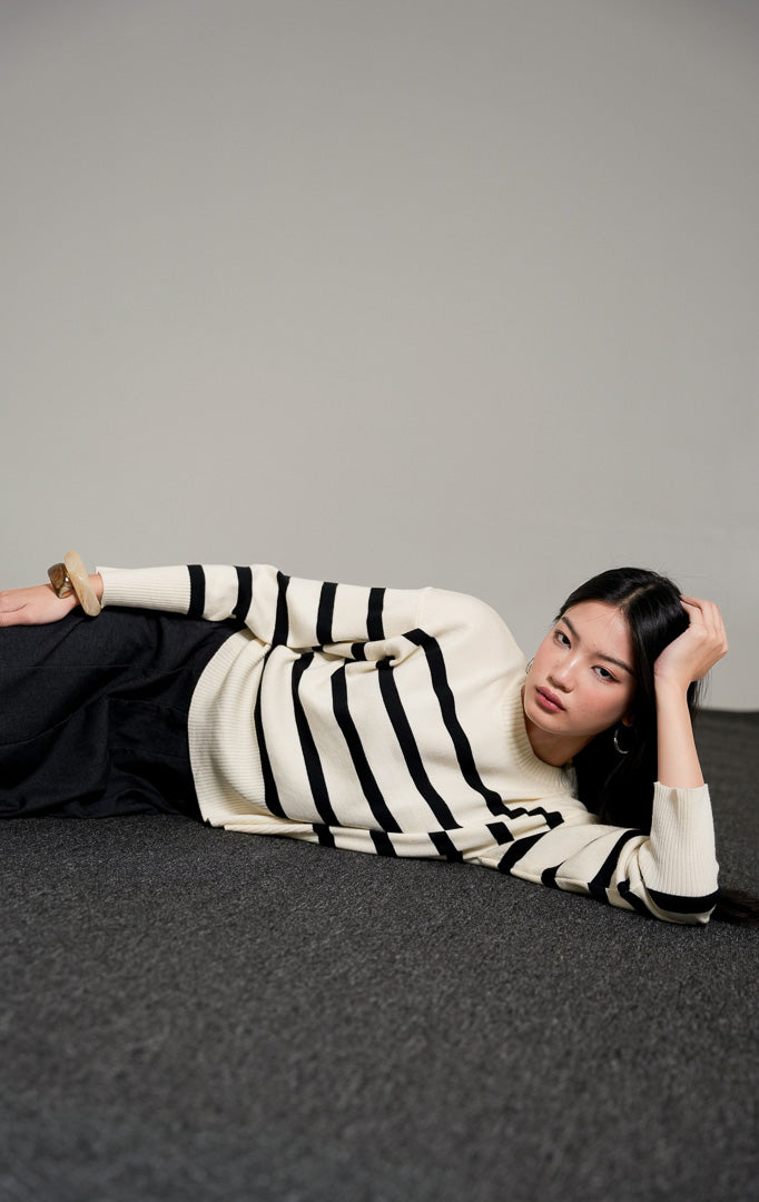 Elbie Striped Knit Sweater lengan panjang warna ivory (gading) dengan motif garis horizontal hitam, dipadukan dengan rok panjang hitam dan sandal bermotif polkadot.
