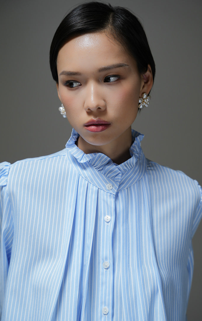 Kemeja wanita Ellenore Top warna biru motif garis salur (stripe) lengan panjang dengan detail kerah ruffle rumbai manis dan kancing depan. 
