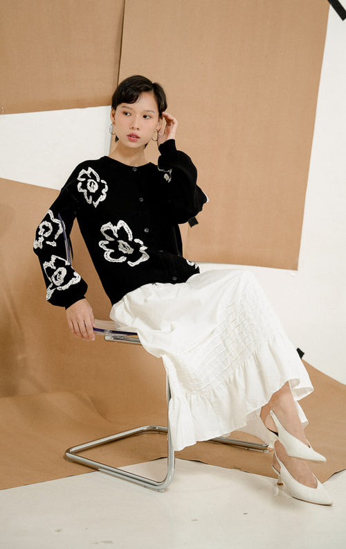 Model berpose duduk mengenakan Emmelie Flower Round-Neck Cardigan Black, produk outerwear cardigan rajut wanita lengan panjang warna hitam dengan motif bunga putih besar. 