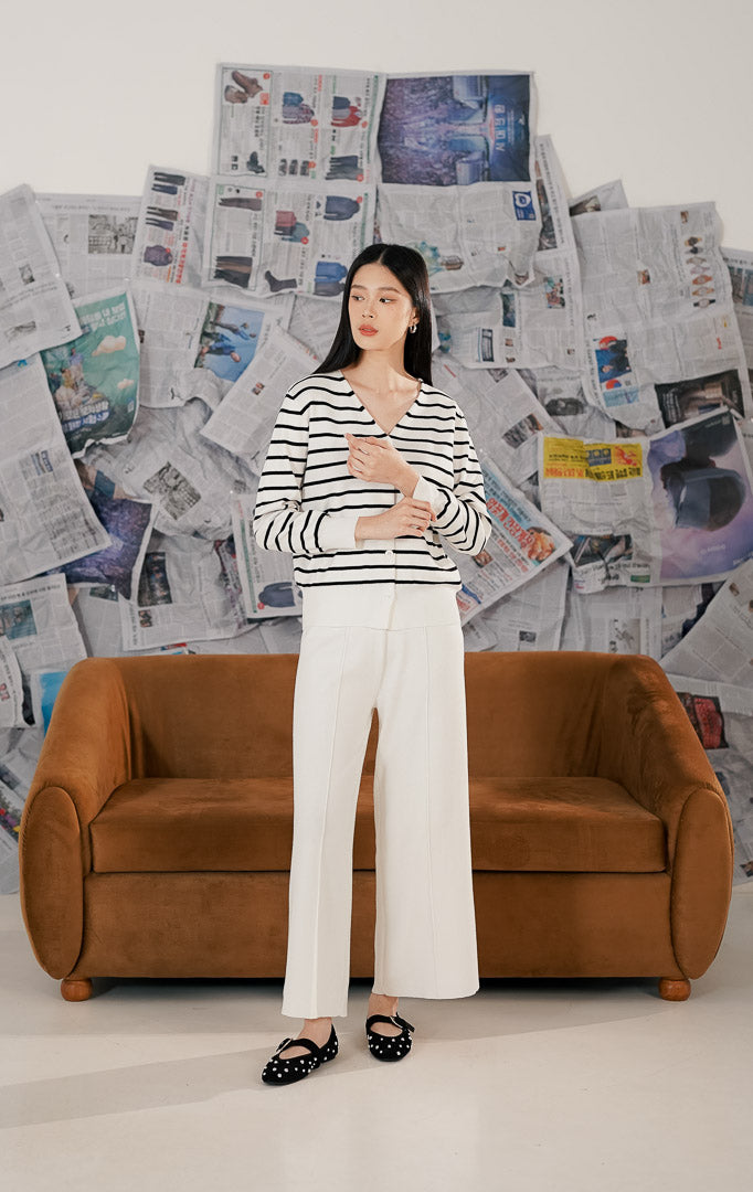 Model wanita mengenakan Esme Cardigan White Striped, sebuah cardigan rajut lengan panjang warna putih dengan motif garis-garis hitam horizontal. Cardigan memiliki potongan leher V-neck, kancing depan warna putih senada, dan bahan rajut lembut.