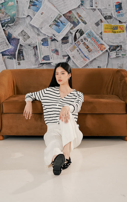 Model wanita mengenakan Esme Cardigan White Striped, sebuah cardigan rajut lengan panjang warna putih dengan motif garis-garis hitam horizontal. Cardigan memiliki potongan leher V-neck, kancing depan warna putih senada, dan bahan rajut lembut.