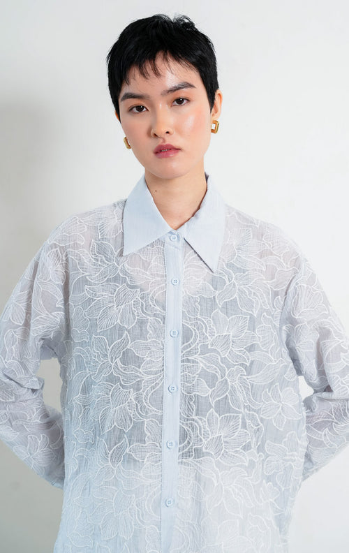 Kemeja wanita lengan panjang warna biru muda (light blue) Everlynne Top berbahan tekstur ringan (lightweight textured fabric) dengan kerah kemeja (collared neckline), bukaan kancing depan, dan detail bordir motif bunga (tonal floral embroidery). 