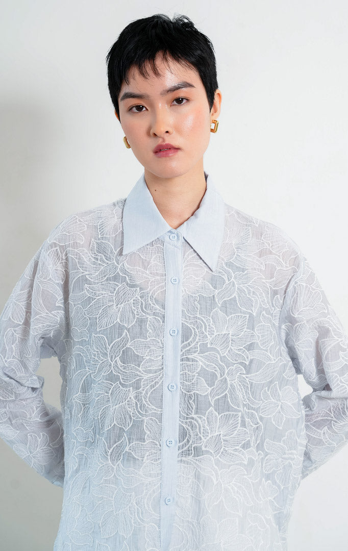 Kemeja wanita lengan panjang warna biru muda (light blue) Everlynne Top berbahan tekstur ringan (lightweight textured fabric) dengan kerah kemeja (collared neckline), bukaan kancing depan, dan detail bordir motif bunga (tonal floral embroidery). 