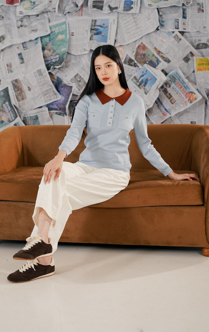 Model wanita mengenakan Eyre Sweater Blue, sebuah atasan rajut lengan panjang warna biru muda (baby blue) dengan kerah wangki warna cokelat bata (terracotta). Baju memiliki detail dua saku dada dan kancing emas, dipadukan dengan celana panjang putih.