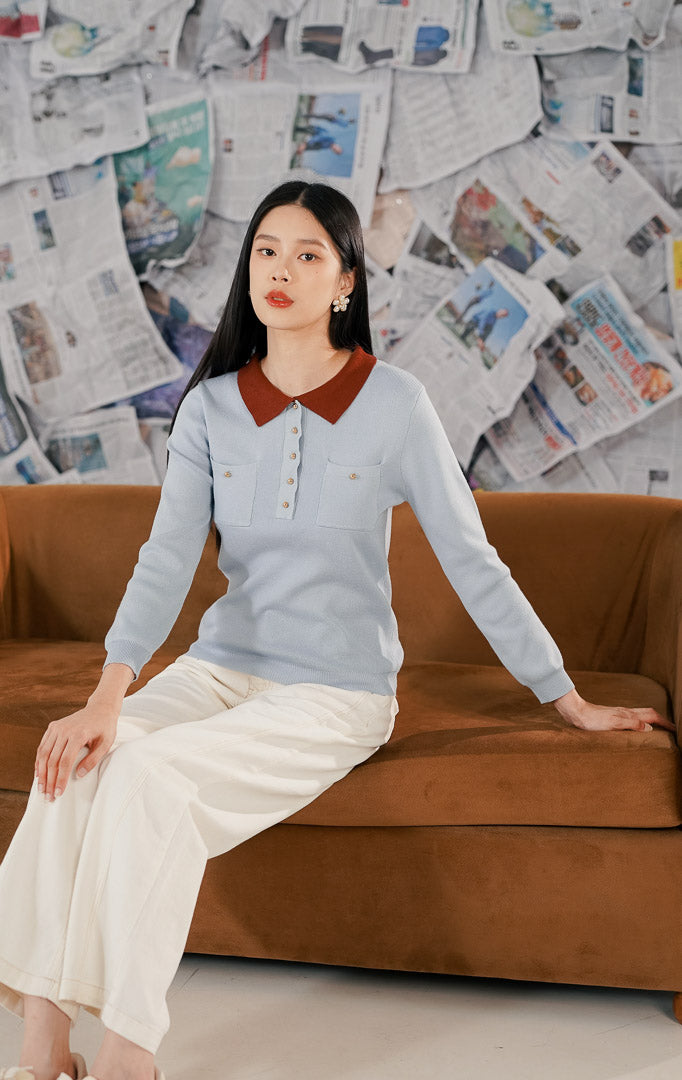 Model wanita mengenakan Eyre Sweater Blue, sebuah atasan rajut lengan panjang warna biru muda (baby blue) dengan kerah wangki warna cokelat bata (terracotta). Baju memiliki detail dua saku dada dan kancing emas, dipadukan dengan celana panjang putih.