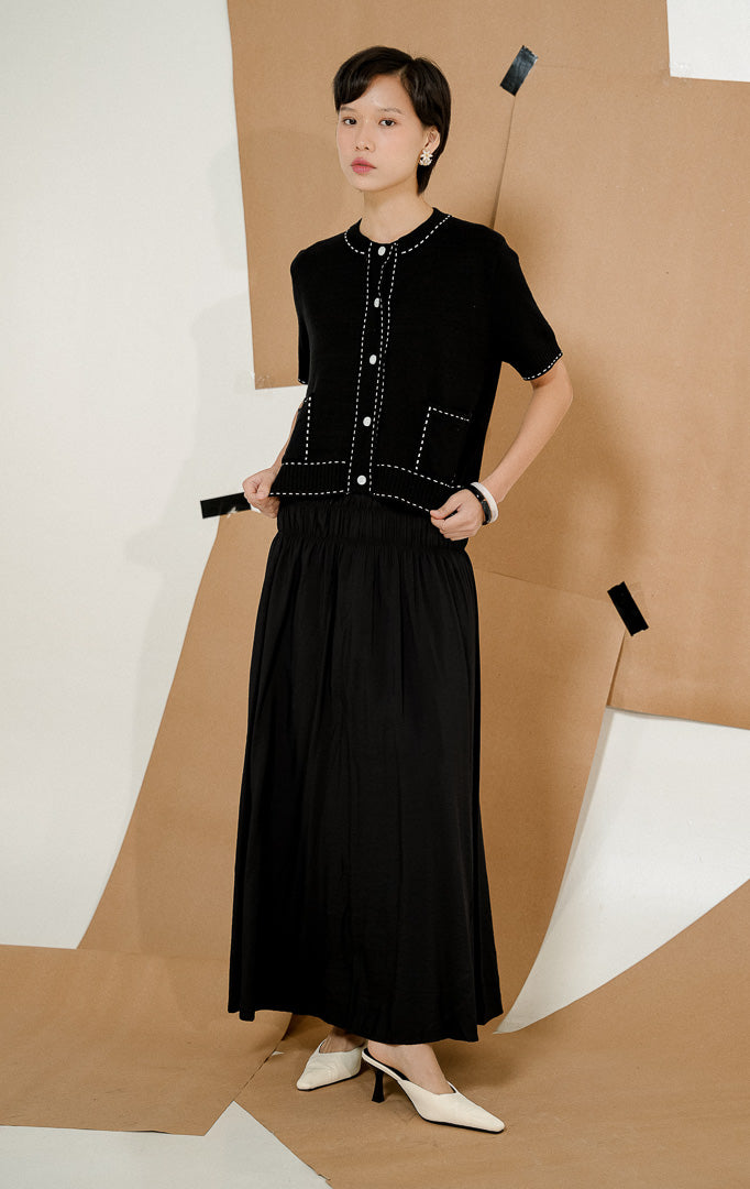 Model berdiri mengenakan Fairmont Stitched-Up Knit Top Black, produk atasan rajut wanita lengan pendek warna hitam dengan detail jahitan kontras putih dan saku depan. 