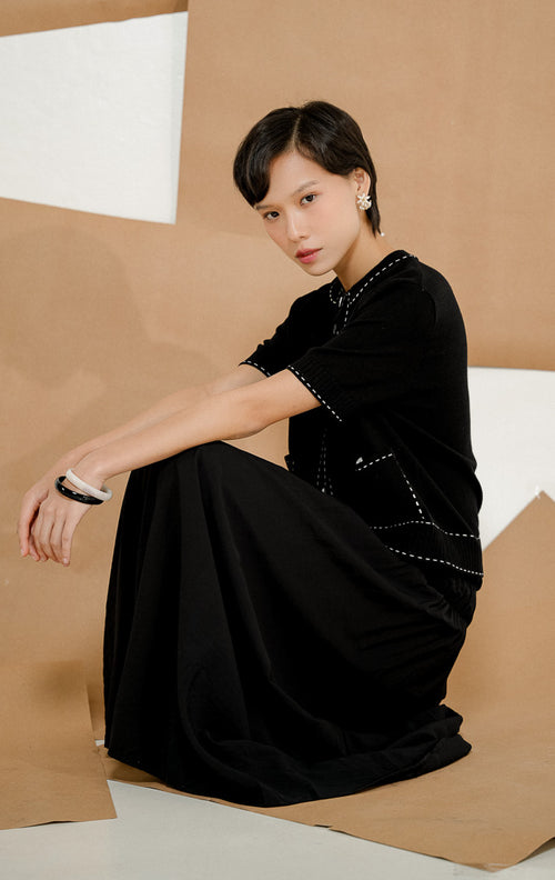 Model berdiri mengenakan Fairmont Stitched-Up Knit Top Black, produk atasan rajut wanita lengan pendek warna hitam dengan detail jahitan kontras putih dan saku depan. 