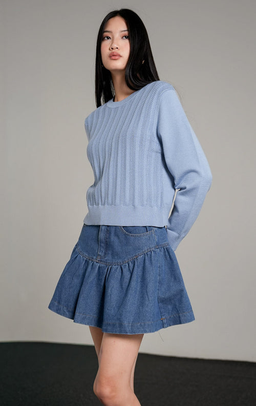 Fifiya Wavy Knit Sweater lengan panjang warna biru muda berbahan rajut dengan pola tekstur garis bergelombang vertikal, dipadukan dengan rok mini denim beraksen ruffle dan sepatu mules putih.