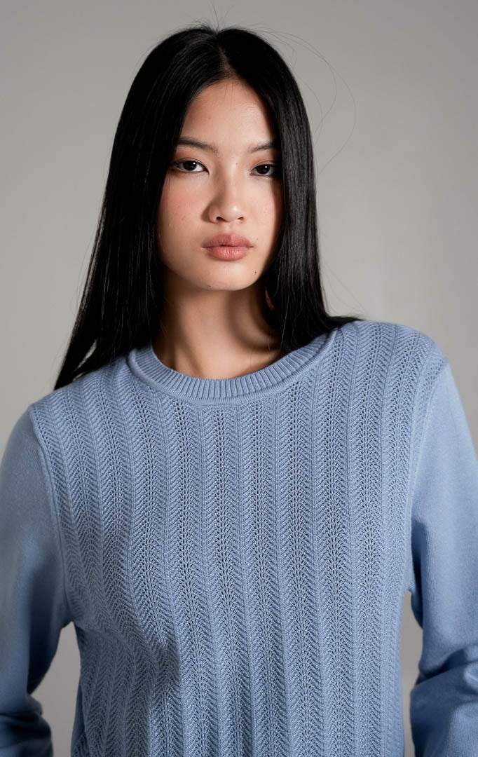 Fifiya Wavy Knit Sweater lengan panjang warna biru muda berbahan rajut dengan pola tekstur garis bergelombang vertikal, dipadukan dengan rok mini denim beraksen ruffle dan sepatu mules putih.