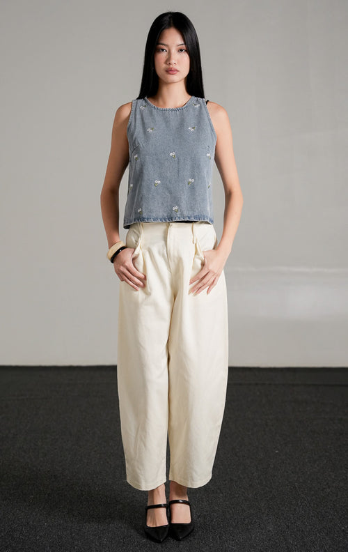 Fleura Top, atasan tanpa lengan warna light denim (biru muda) dengan detail bordir bunga kecil tersebar, dipadukan dengan celana panjang gading berpotongan longgar dan sepatu hak hitam.
