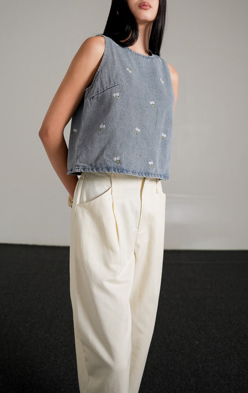 Fleura Top, atasan tanpa lengan warna light denim (biru muda) dengan detail bordir bunga kecil tersebar, dipadukan dengan celana panjang gading berpotongan longgar dan sepatu hak hitam.