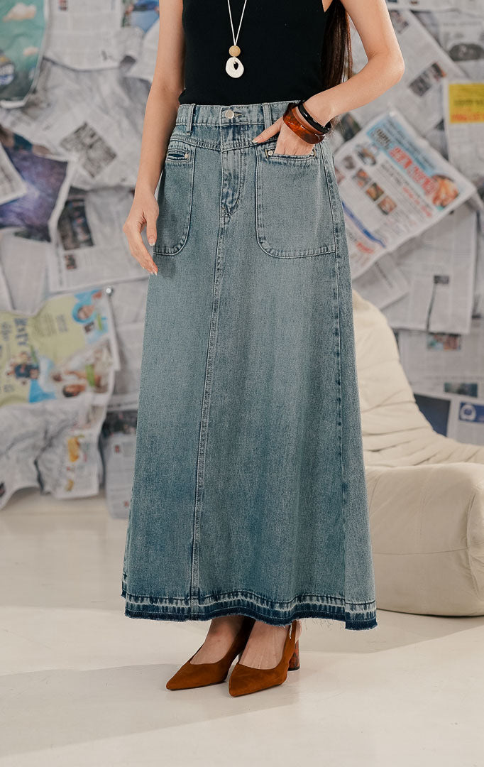 Model wanita mengenakan Franca Skirt Denim, sebuah rok jeans panjang (maxi denim skirt) warna biru medium wash. Rok memiliki dua saku depan besar (patch pockets), pinggang karet, dan detail ujung bawah yang tidak dijahit (raw hem), dipadukan dengan tank top hitam.