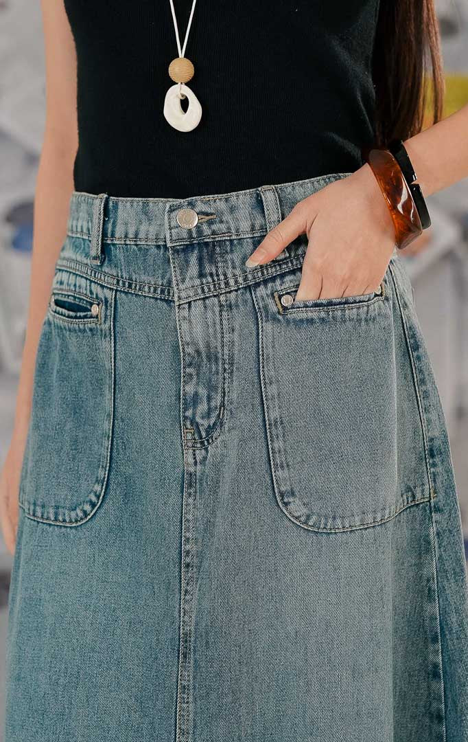 Model wanita mengenakan Franca Skirt Denim, sebuah rok jeans panjang (maxi denim skirt) warna biru medium wash. Rok memiliki dua saku depan besar (patch pockets), pinggang karet, dan detail ujung bawah yang tidak dijahit (raw hem), dipadukan dengan tank top hitam.