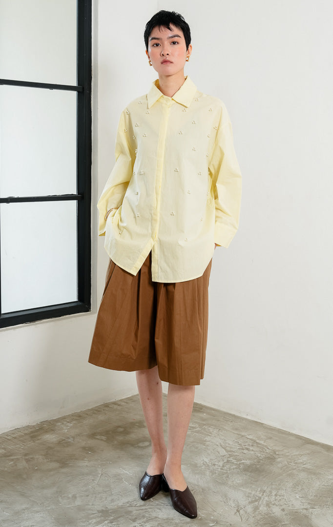 Kemeja wanita lengan panjang warna kuning muda (butter yellow) Freesia Top dengan kerah klasik (classic collar), bukaan kancing depan tertutup, dan hiasan manik mutiara motif bunga (pearl bead embellishments).