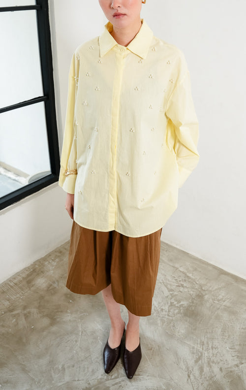 Kemeja wanita lengan panjang warna kuning muda (butter yellow) Freesia Top dengan kerah klasik (classic collar), bukaan kancing depan tertutup, dan hiasan manik mutiara motif bunga (pearl bead embellishments).