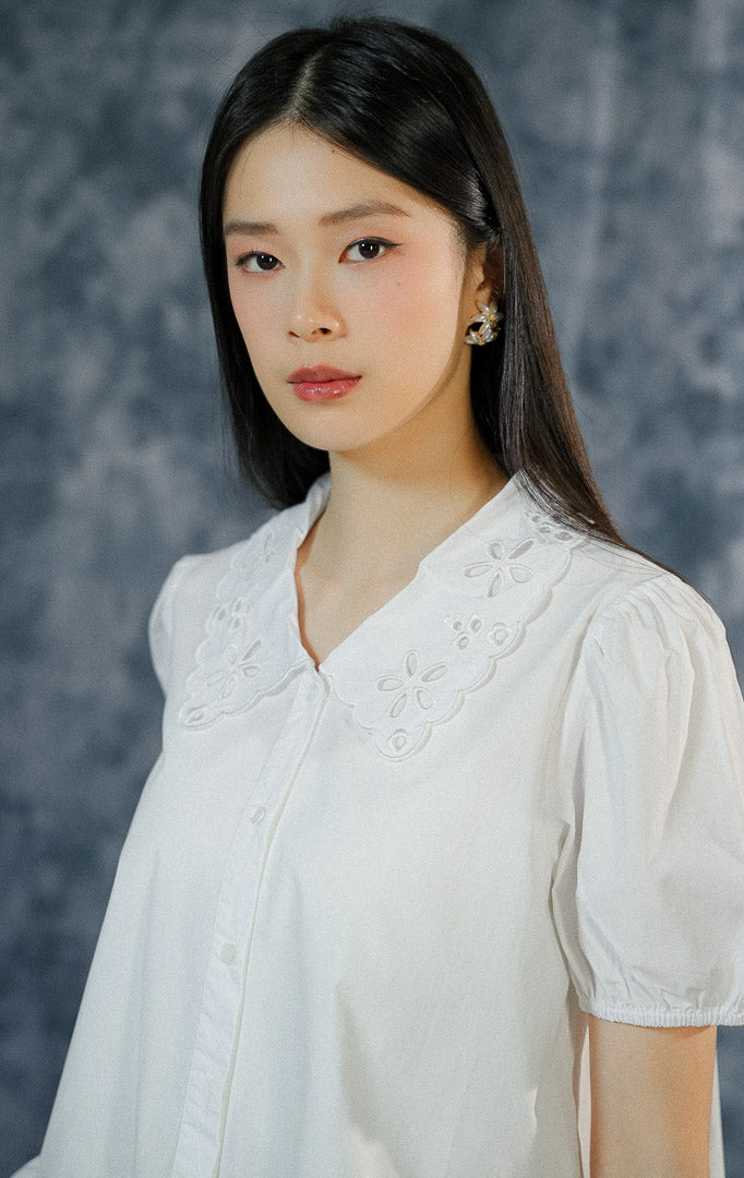 Atasan kemeja lengan pendek wanita Ginette warna putih polos dengan detail kerah scallop bersulam, ujung lengan karet puff, dan aksen ruffled hem bawah.