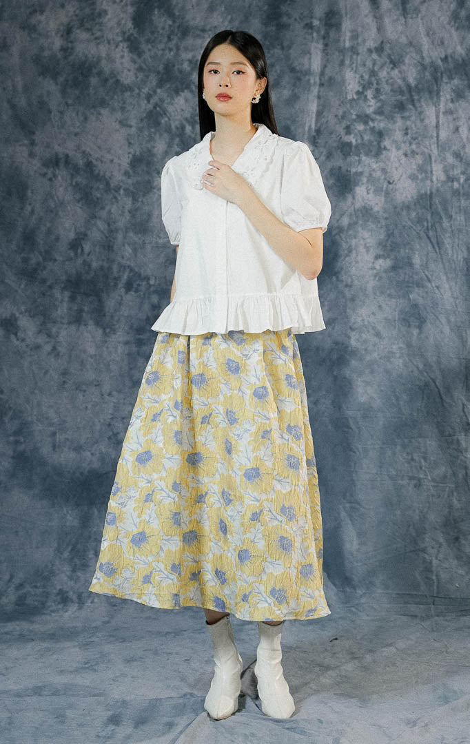 Atasan kemeja lengan pendek wanita Ginette warna putih polos dengan detail kerah scallop bersulam, ujung lengan karet puff, dan aksen ruffled hem bawah.
