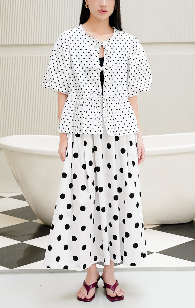 Baju atasan wanita Giselle Top warna putih motif polkadot hitam dengan detail tali ikatan depan (tie-front), lengan balon puff sleeve, dan potongan peplum mekar.