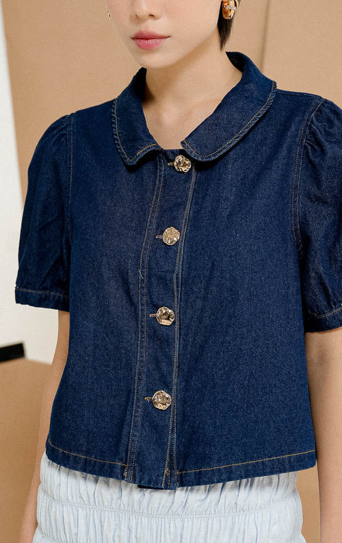 Model berdiri mengenakan kemeja Hanson Peter Pan Collar Blouse in Dark Denim, produk atasan denim wanita lengan pendek dengan kerah bulat dan detail kancing emas. 