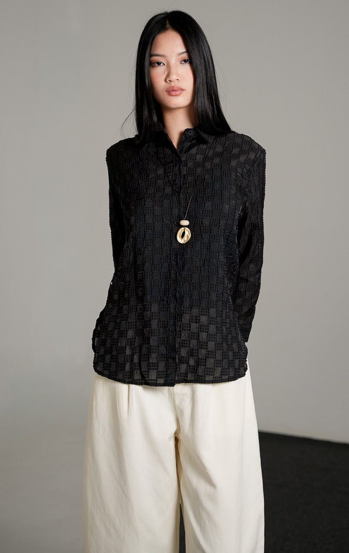 kemeja lengan panjang Hazel Top warna hitam berbahan katun dengan detail bordir pola kotak bertekstur, dipadukan dengan celana panjang broken white dan sepatu heels hitam.
