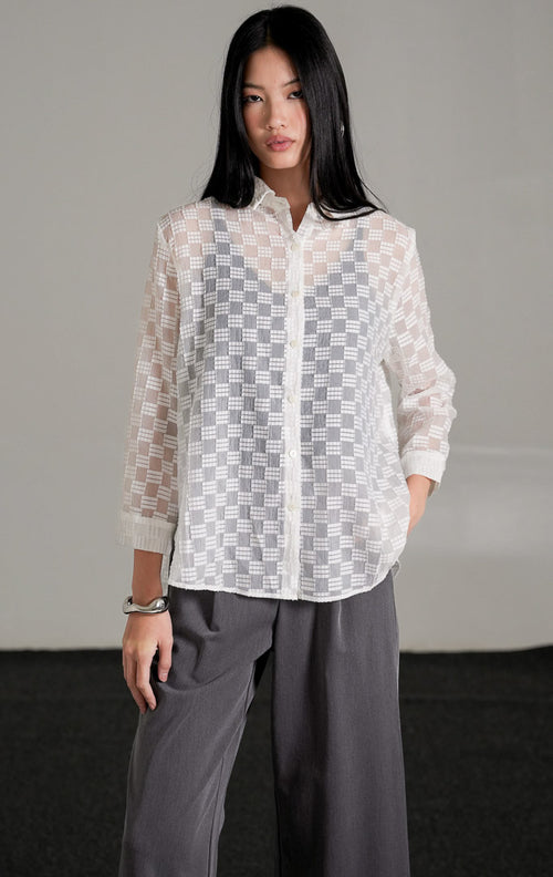 Hazel Top warna putih dengan detail bordir pola kotak semi-transparan yang memperlihatkan inner hitam di baliknya, dipadukan dengan celana kulot abu-abu dan sepatu heels putih.