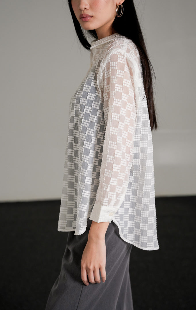  Hazel Top warna putih dengan detail bordir pola kotak semi-transparan yang memperlihatkan inner hitam di baliknya, dipadukan dengan celana kulot abu-abu dan sepatu heels putih.