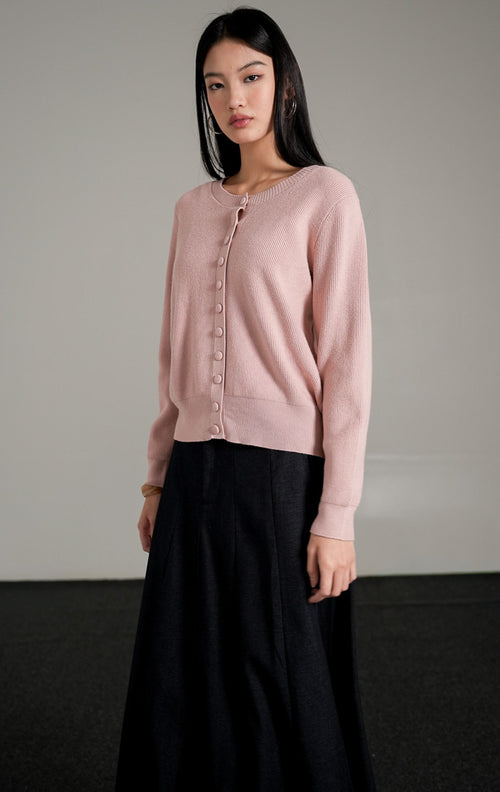 Hermione Knit Cardigan lengan panjang berwarna dusty pink dengan kancing depan tertutup, dipadukan dengan rok midi A-line hitam dan sepatu heels putih.
