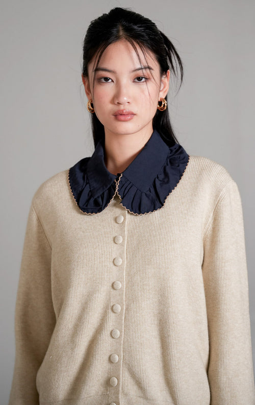 Hermione Knit Cardigan lengan panjang berwarna ivory (gading) dengan kancing depan tertutup, dipadukan dengan celana panjang bahan warna cokelat dan sepatu heels putih.
