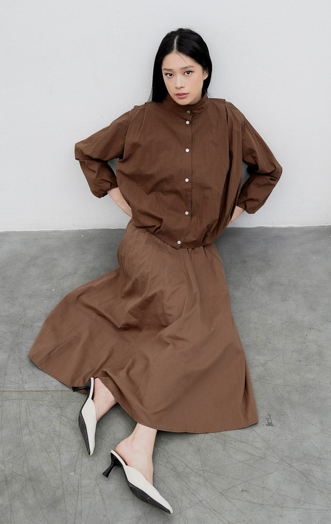 Jaket wanita warna cokelat tua (dark brown) Honoria Jacket berbahan campuran crinkled cotton-nylon dengan kerah tinggi (high-neck), bukaan kancing jepret (snap-button closure), dan saku fungsional.