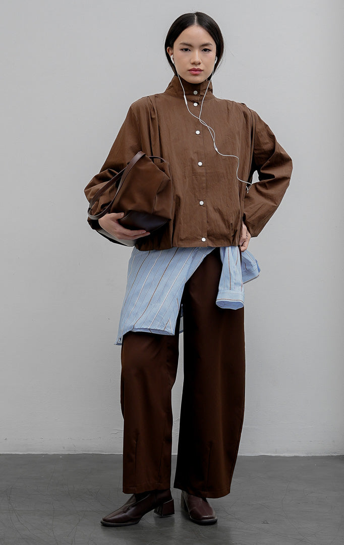 Jaket wanita warna cokelat tua (dark brown) Honoria Jacket berbahan campuran crinkled cotton-nylon dengan kerah tinggi (high-neck), bukaan kancing jepret (snap-button closure), dan saku fungsional.