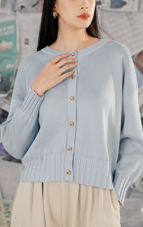 Model wanita mengenakan Hudson Cardigan Blue, sebuah cardigan rajut (knitwear) lengan panjang warna biru muda pastel dengan kerah bulat dan detail kancing depan berwarna emas.