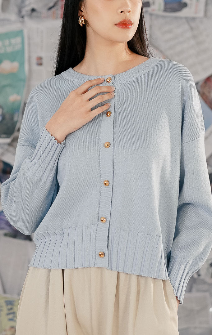 Model wanita mengenakan Hudson Cardigan Blue, sebuah cardigan rajut (knitwear) lengan panjang warna biru muda pastel dengan kerah bulat dan detail kancing depan berwarna emas.