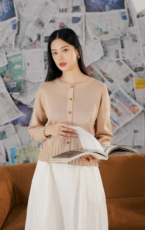 Model wanita mengenakan Hudson Cardigan Khaki, sebuah cardigan rajut (knitwear) lengan panjang warna krem kecokelatan dengan kerah bulat dan kancing depan emas, sambil memegang majalah.