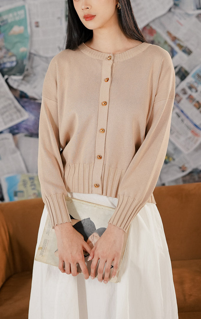 Model wanita mengenakan Hudson Cardigan Khaki, sebuah cardigan rajut (knitwear) lengan panjang warna krem kecokelatan dengan kerah bulat dan kancing depan emas, sambil memegang majalah.