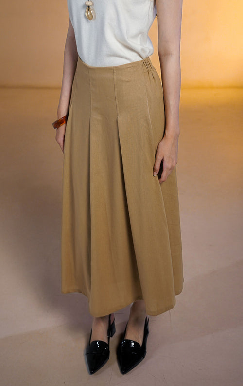 Model wanita mengenakan Hudson Skirt Brown, rok midi potongan A-line warna cokelat muda dengan detail lipatan halus (semi-pleated) dan pinggang elastis