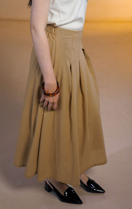 Model wanita mengenakan Hudson Skirt Brown, rok midi potongan A-line warna cokelat muda dengan detail lipatan halus (semi-pleated) dan pinggang elastis