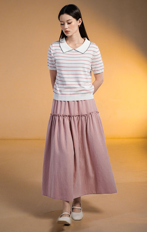 Model wanita mengenakan Hunter Outer Pink, atasan rajut lengan pendek motif garis-garis pink dan putih dengan kerah putih bertepi hitam tanpa kancing, dipadukan dengan rok ruffle panjang warna pink dusty