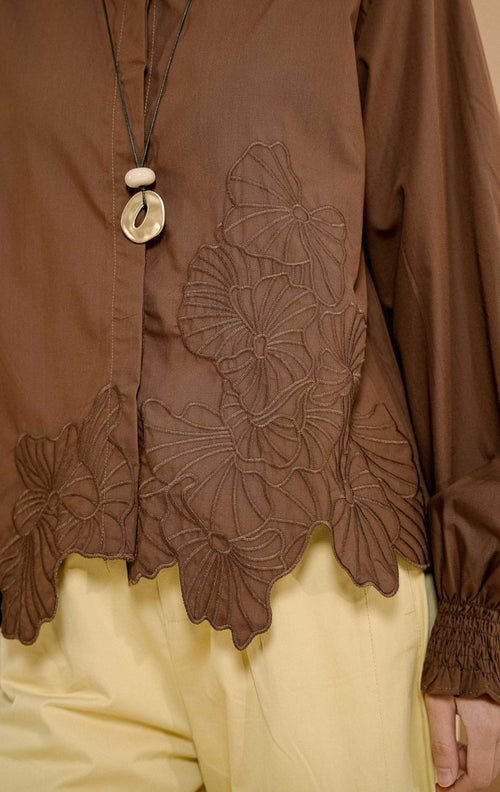 Model mengenakan Iridessa Embroidered Button-Up Shirt Dark Brown, kemeja katun wanita lengan panjang warna cokelat tua dengan detail bordir bunga dan ujung keliman bergelombang. 